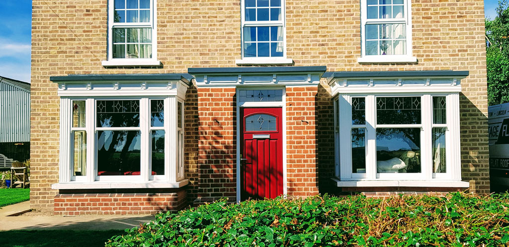 A charming brick house features a striking red front door and large bay windows on either side. Lush greenery surrounds the base, adding to the inviting atmosphere.