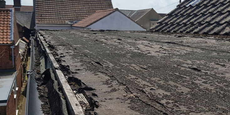 The image depicts a weathered and worn flat roof, showing signs of deterioration with cracked and peeling surfaces. Nearby rooftops are visible in the background, indicating an urban setting.