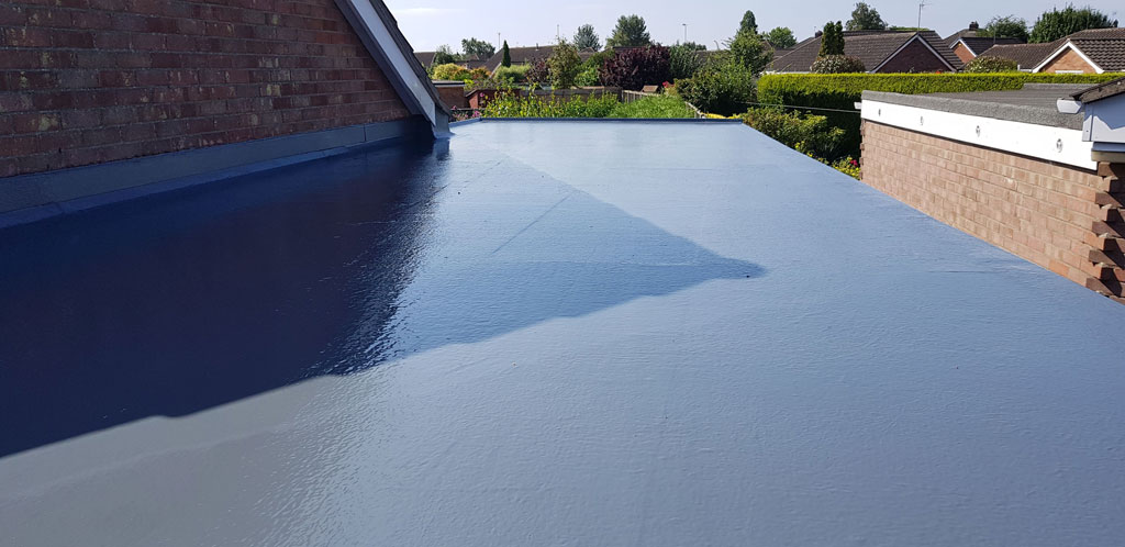 A flat roof is shown with a smooth, freshly applied gray coating. In the background, residential houses and greenery are visible under a clear blue sky.