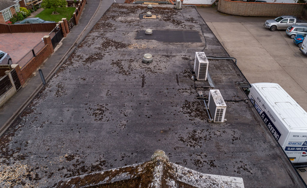 A flat roof is visible, showing signs of wear with patches of dirt and debris scattered across the surface. Air conditioning units are positioned along the edge, with a view of a nearby parking area and residential buildings in the background.