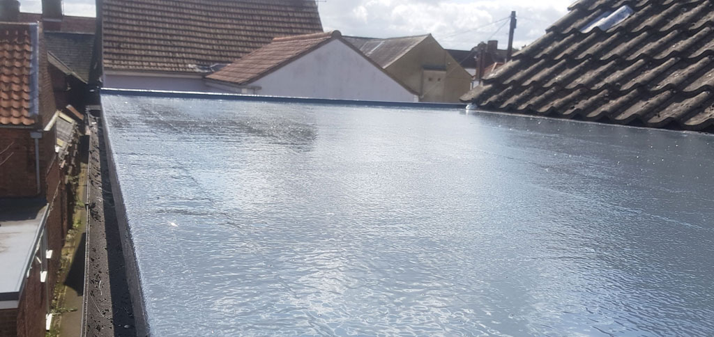 A flat roof with a shiny, reflective surface is visible, surrounded by various rooftops in the background. The scene is bright, indicating a sunny day with some clouds.