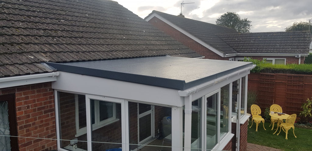 A flat roof extends over a conservatory, featuring a smooth, dark surface. In the background, there are yellow garden chairs and a well-maintained garden area.