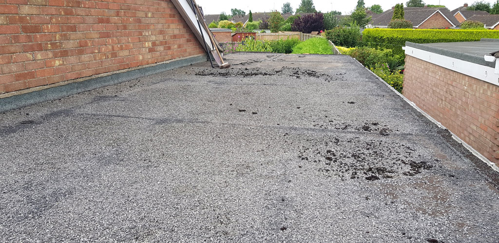 A flat rooftop is shown, featuring a surface with scattered debris and patches of dark material. In the background, residential houses and greenery are visible.