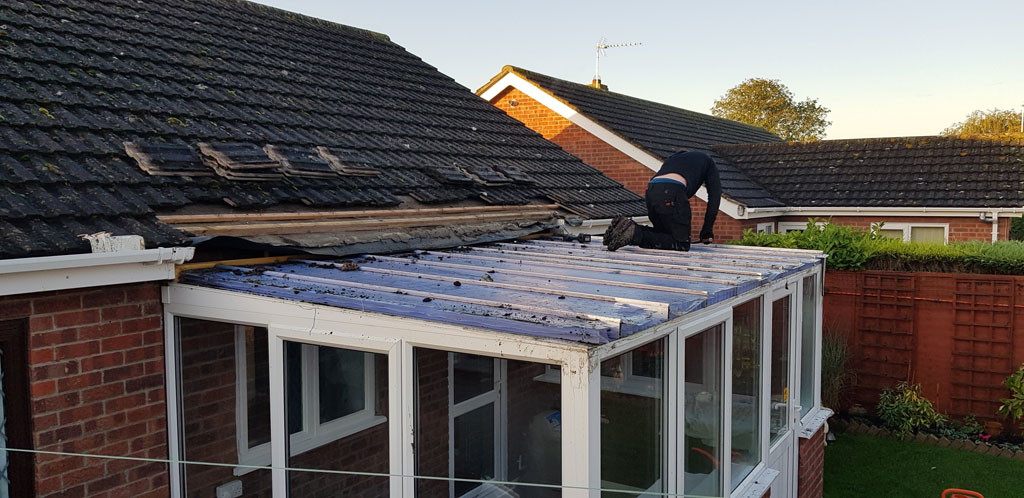 A person is working on the roof of a conservatory, repairing or replacing the roofing material. The surrounding area includes a brick house and a well-maintained garden.