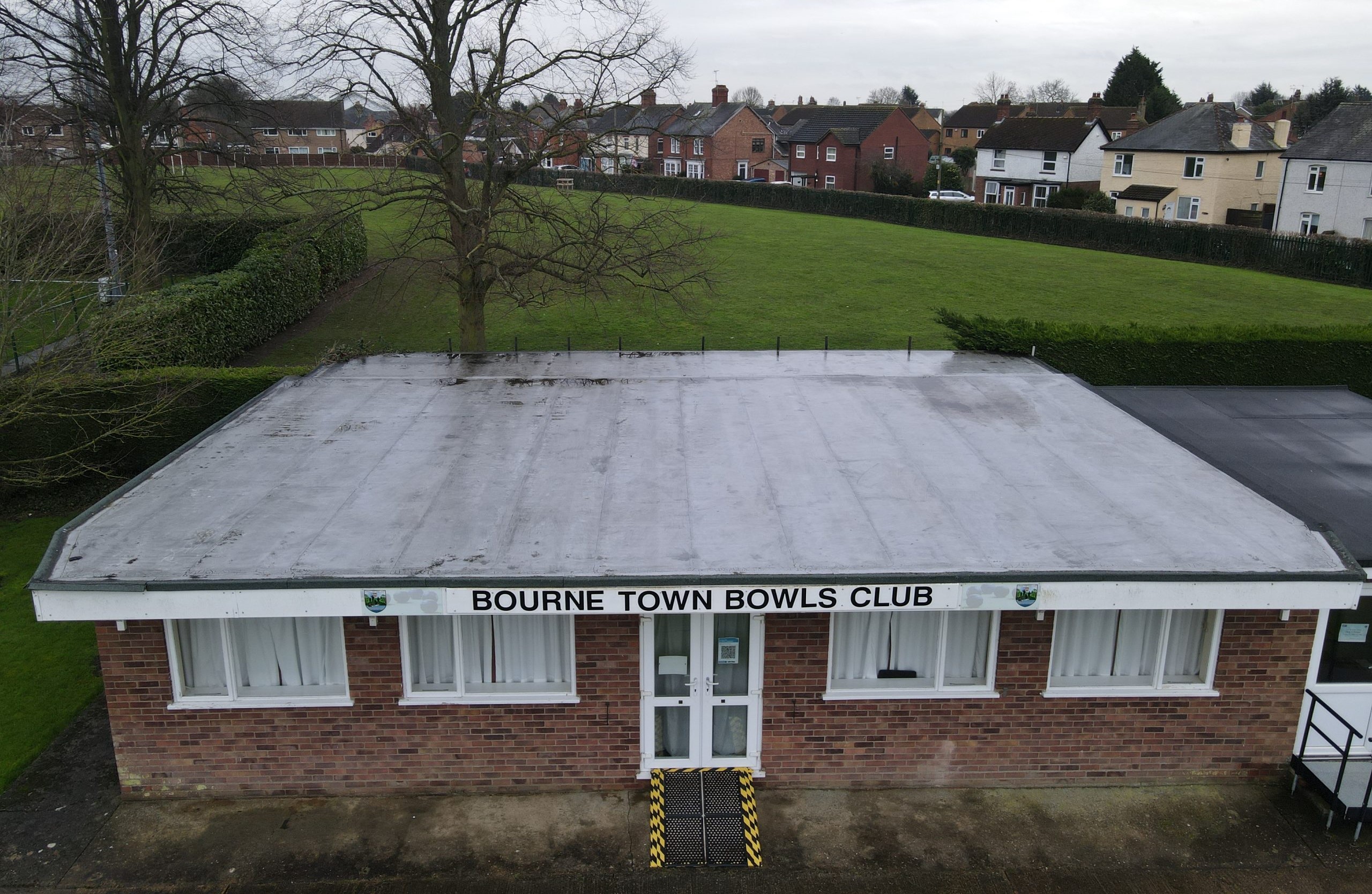 A brick building with a flat roof is labeled "Bourne Town Bowls Club." It is surrounded by green grass and trees, with residential houses visible in the background.