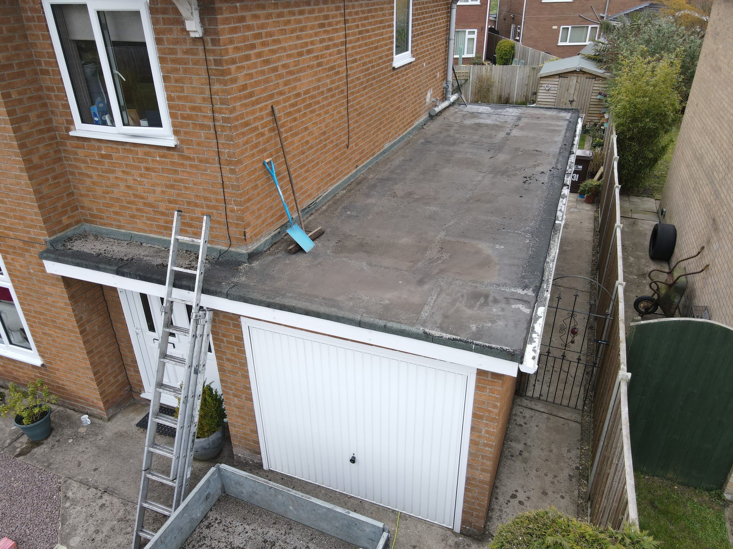 A flat roof is visible on a brick building, with a ladder leaning against the side. Nearby, there are various outdoor items, including a shovel and a gate leading to a side path.