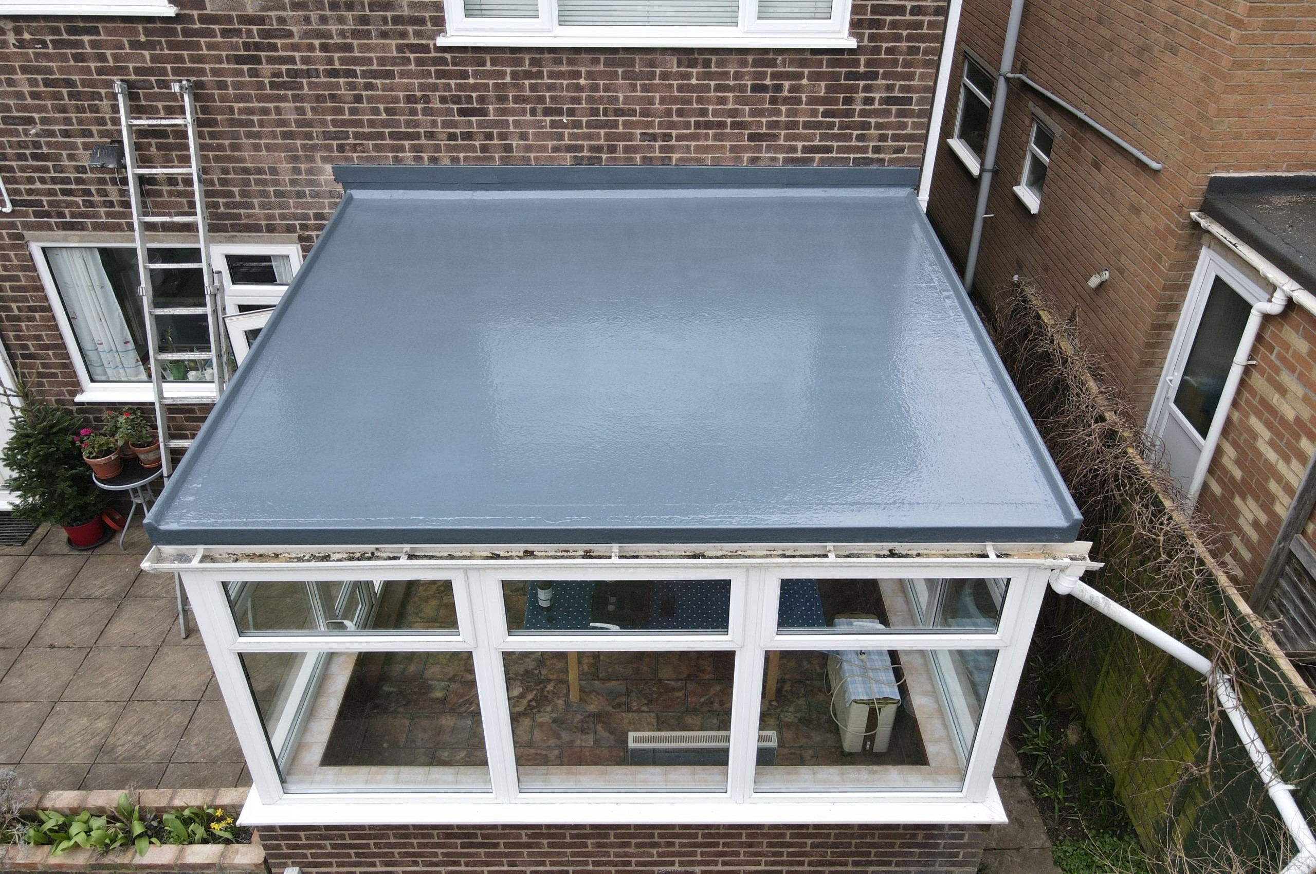 A flat, freshly painted gray roof is visible above a conservatory, with a view of the surrounding brick walls and a ladder leaning against the house. Inside the conservatory, a table and chairs can be seen through the large windows.