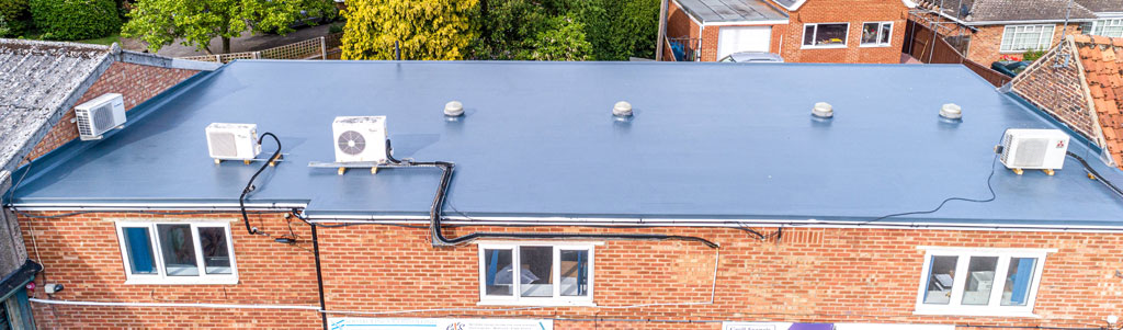 A flat roof is visible, featuring several air conditioning units and ventilation pipes. Surrounding greenery and neighboring buildings can be seen in the background.