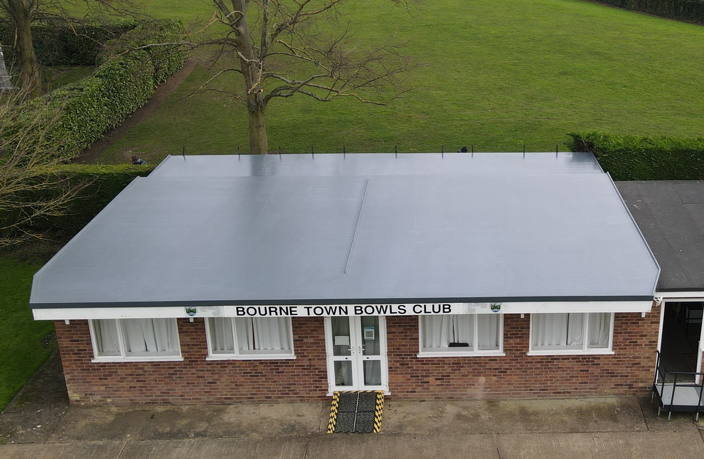 A building with a flat gray roof and a sign reading "BOURNE TOWN BOWLS CLUB" is visible. The structure features large windows and is surrounded by green grass and hedges.