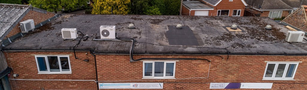 The image depicts the rooftop of a brick building featuring several air conditioning units and ventilation systems. The surface shows signs of wear, with patches of dirt and debris, and there are multiple windows visible along the side of the building.