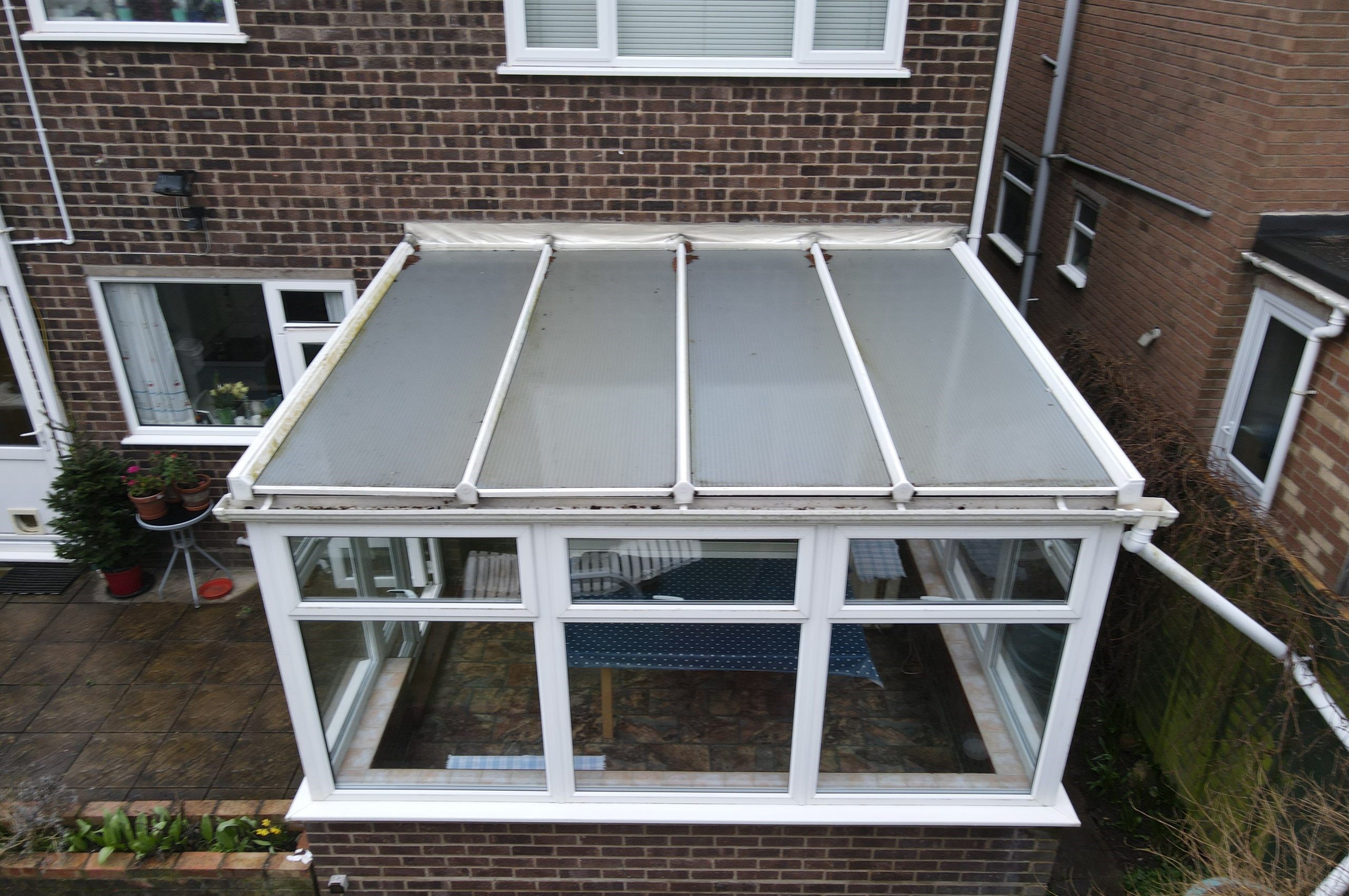 A conservatory with a sloped roof is visible, featuring white frames and glass panels. Inside, there are wooden furniture pieces and a blue tablecloth, with a brick wall and potted plants outside.